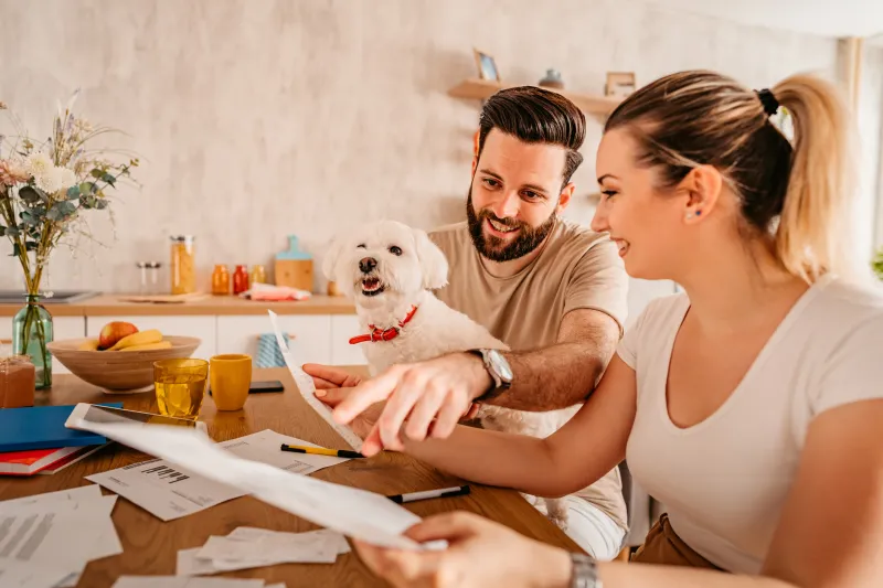 A couple sitting at a computer with paperwork. The man has a small white dog on his lap.