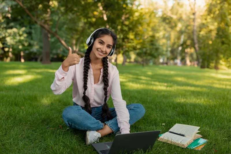 Young girl sitting in the grass with her laptop giving a thumbs up.