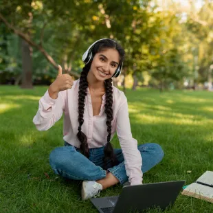 Young girl sitting in the grass with her laptop giving a thumbs up.